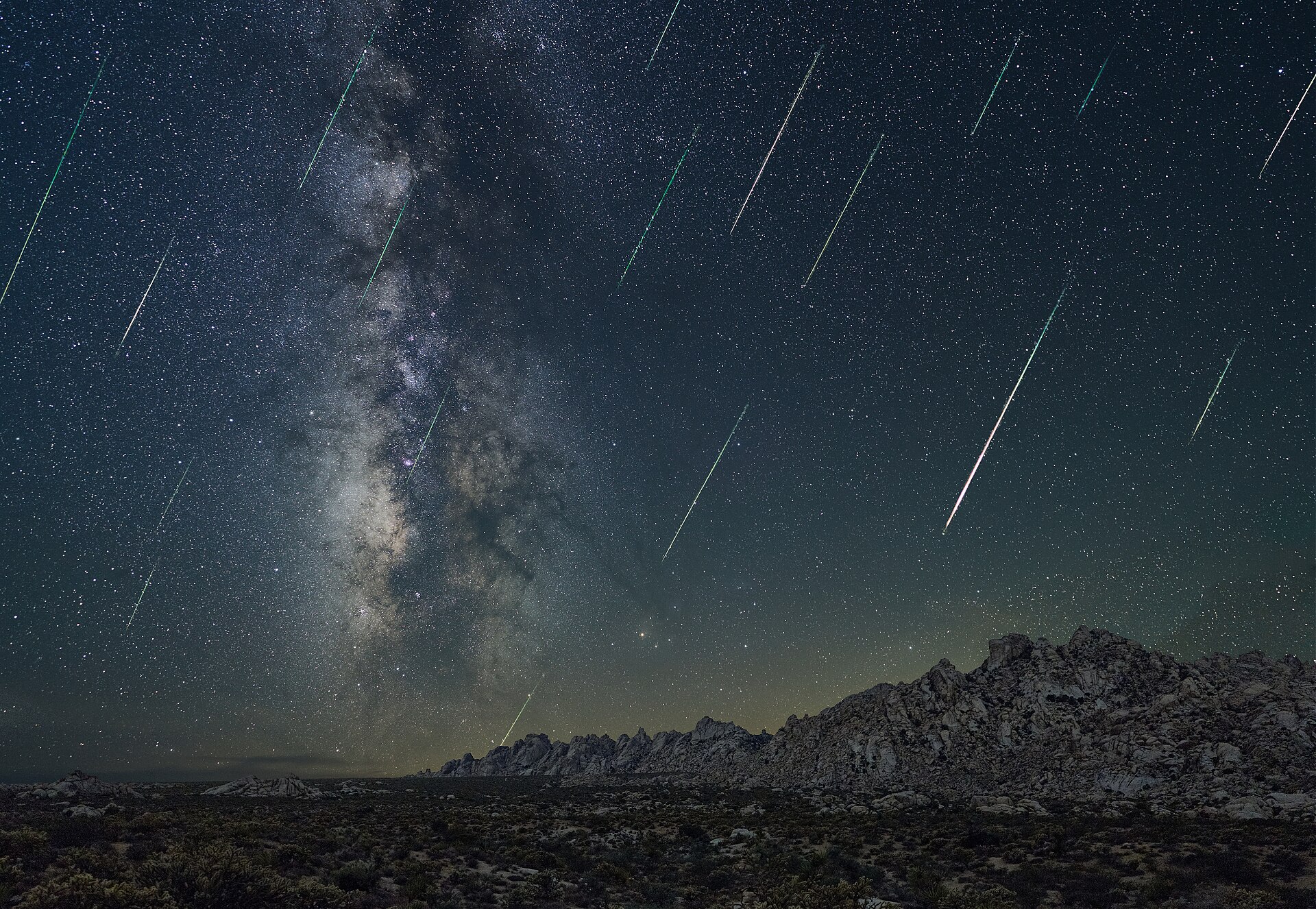A landscape with desert mountains in. In the background is the milky way and shooting stars all over in the view.