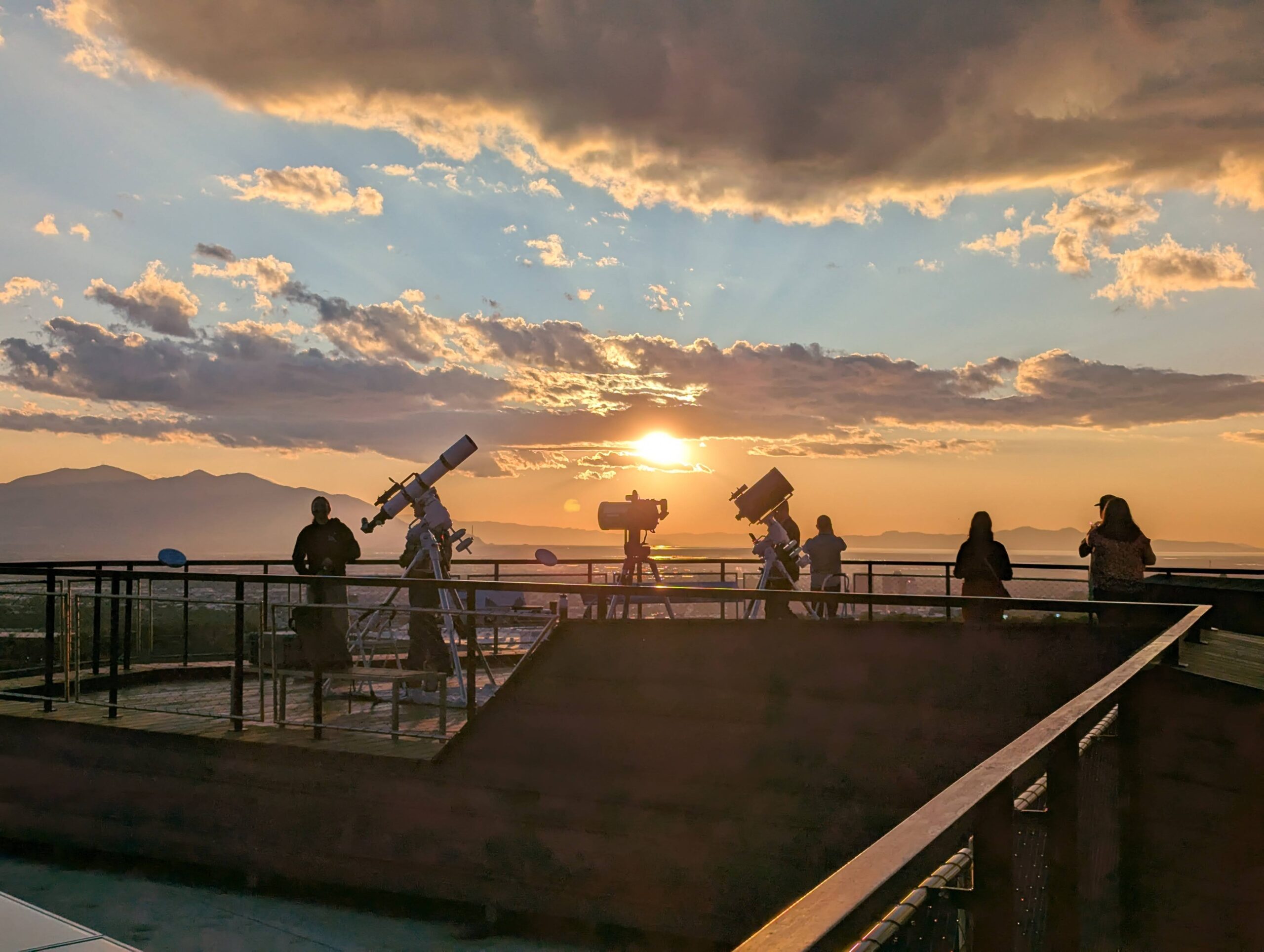 Three Telescopes and few people are on a deck lookin at the sunset above distant mountains. The sun is near clouds in the sky.